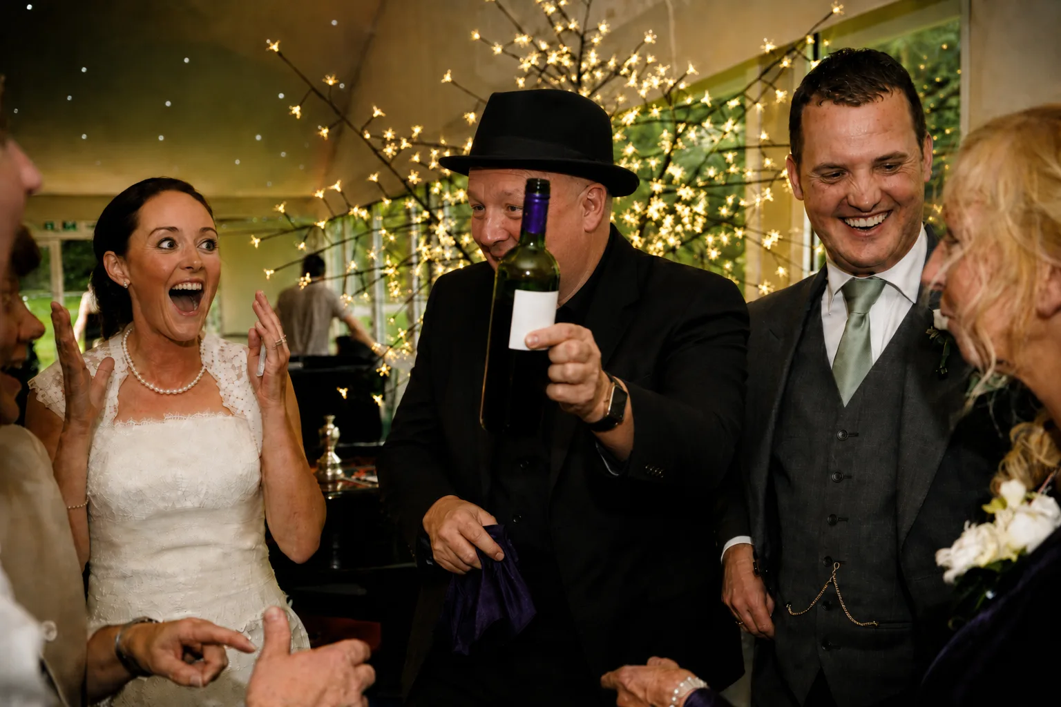 A bride reacts with delight as Clive performs a close-up reveal at a wedding reception while the groom and family watch and laugh.